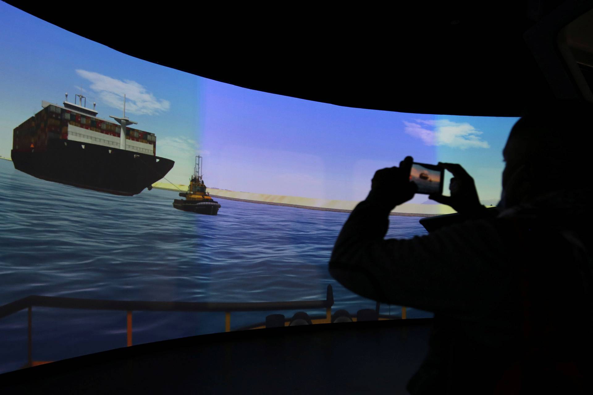 A man records a video as he watches a screen displaying a simulation of a ship being towed by a tug, at SCA Maritime Training and Simulation Centre, in Ismailia