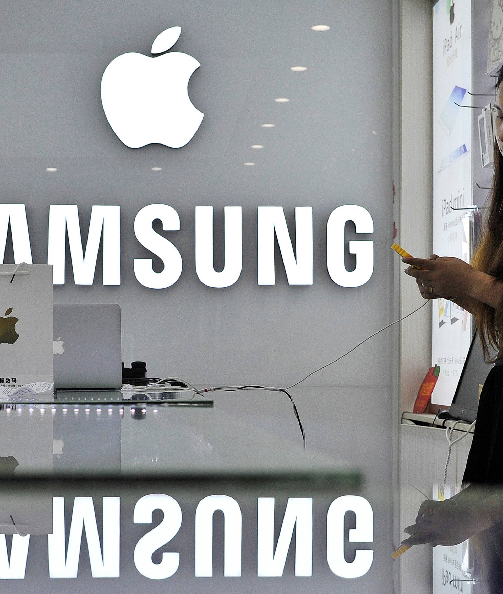 FILE PHOTO: A sales assistant uses her mobile phone next to the company logos of Apple and Samsung at a store in Hefei