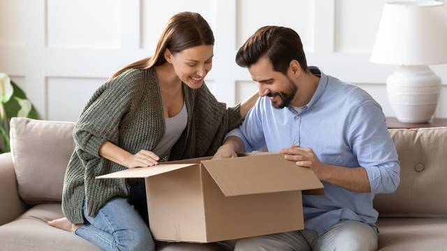 Happy,Curious,Couple,Unpacking,Parcel,At,Home,,Sitting,On,Couch,