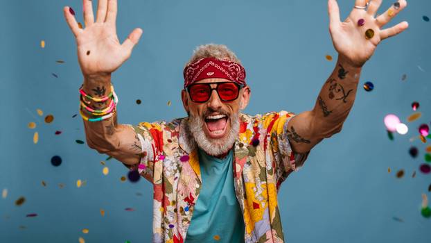 Joyful senior man in funky shirt and bandana throwing colorful confetti on blue background