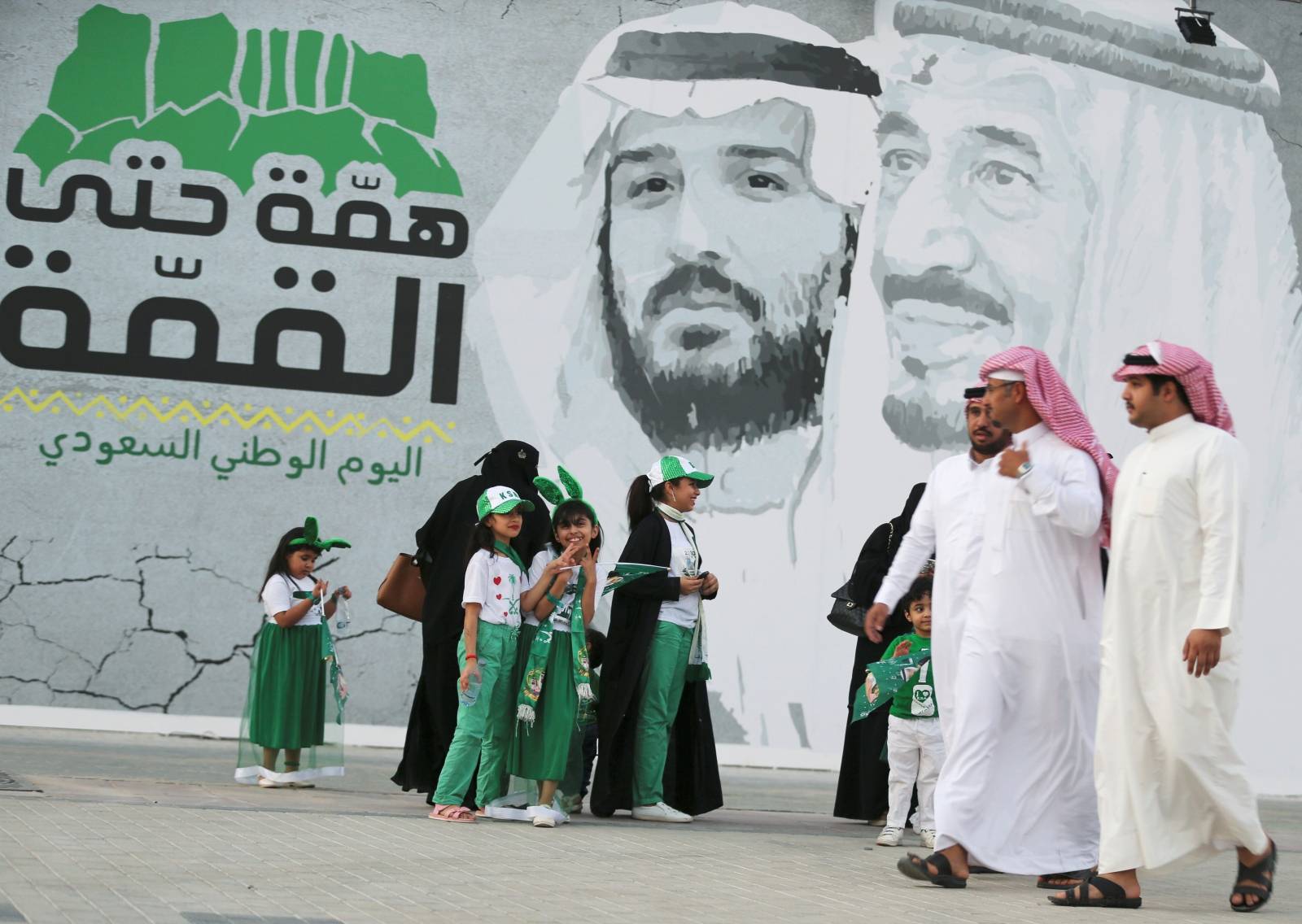 Saudi people walk past a poster depicting Saudi Arabia's King Salman bin Abdulaziz and Crown Prince of Saudi Arabia Mohammad bin Salman during the 89th annual National Day of Saudi Arabia in Riyadh