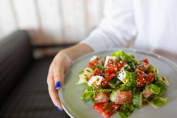 The young female waiter holding in her hands the plate with the Greek salad.