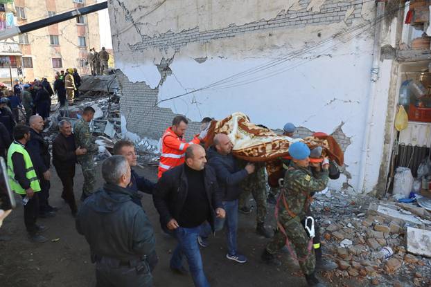 Military and emergency personnel carry an injured man on a stretcher near a damaged building in