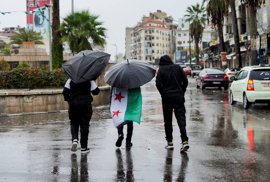 A person with the Syrian flag draped over his shoulders with an umbrella walks with others as Syrians mark the first anniversary of Bashar al-Assad's fall, in Latakia