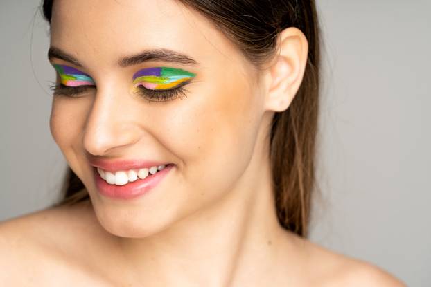 happy teen model with colorful makeup smiling isolated on grey
