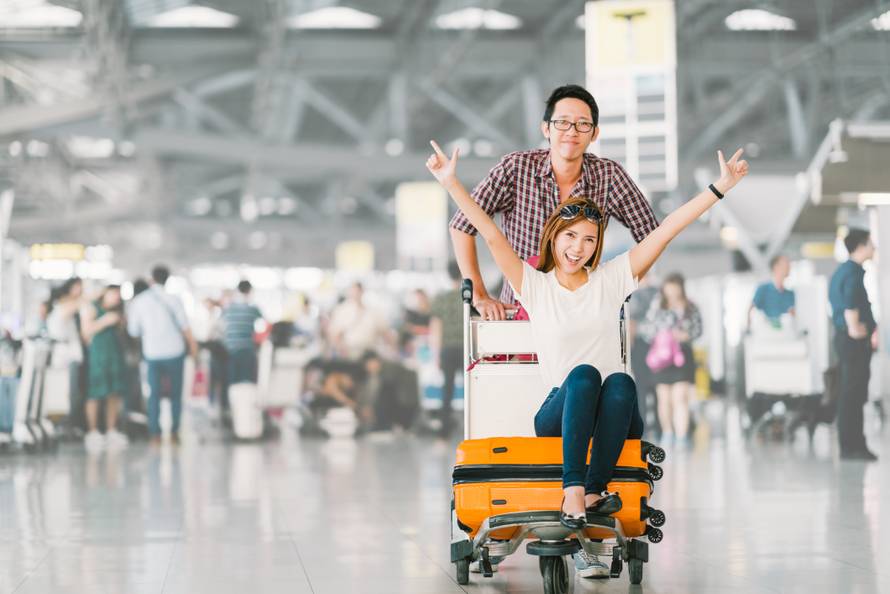 Young Asian tourist couple happy and excited together for the trip, girlfriend sitting and cheering on baggage trolley or luggage cart. Holiday vacation travelling abroad concept, with copy space