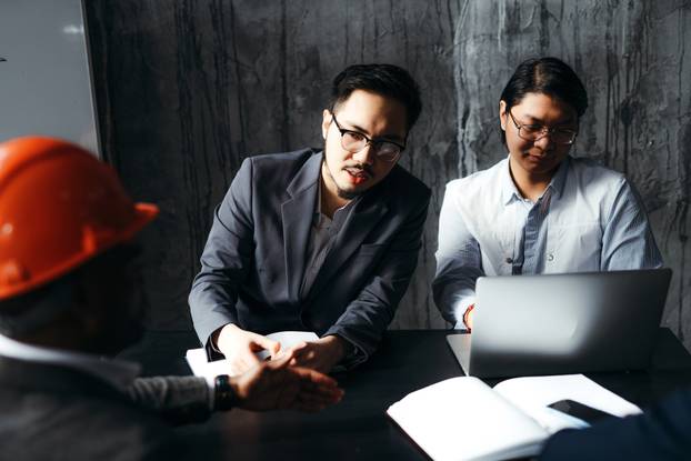 Two korean workers in suits are sitting at a desk working at a computer and communicating with an african american man in a helmet