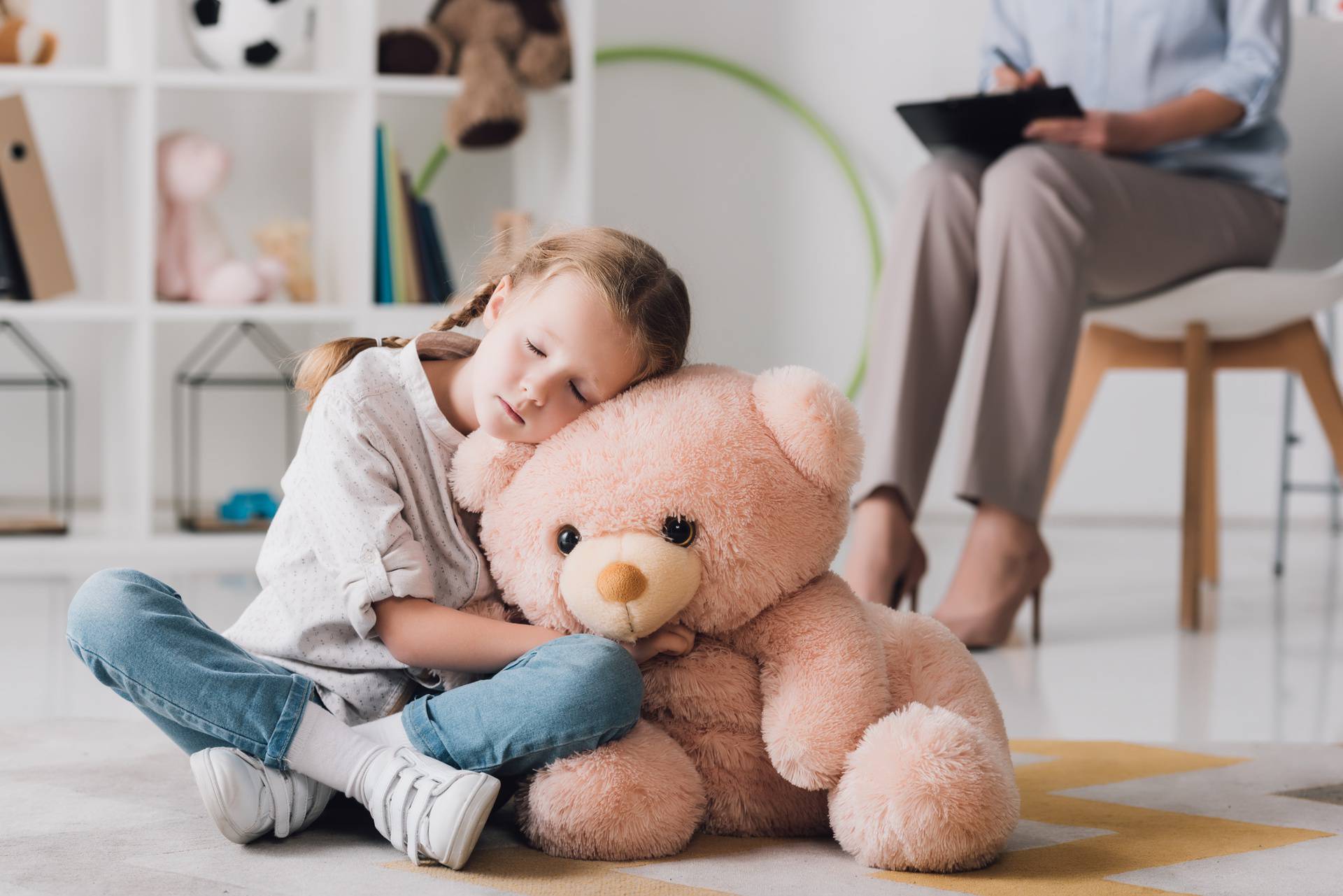 lonely little child with teddy bear sitting on floor with psychologist sitting on background