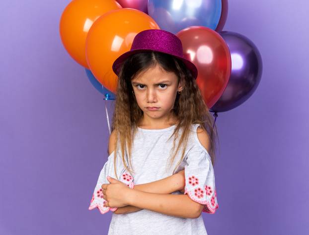 offended little caucasian girl with violet party hat standing with crossed arms in front of helium balloons isolated on purple background with copy space