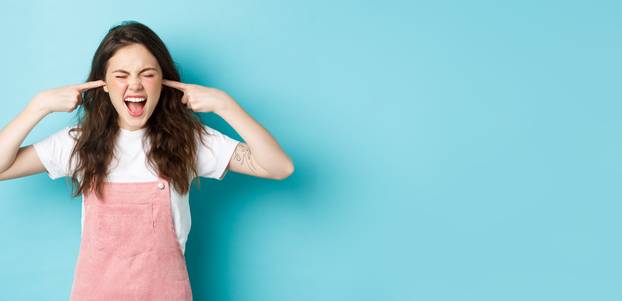 Annoyed young brunette woman screaming and shutting ears, blocking annoying sound, ignoring someone, hear awful noise, standing against blue background