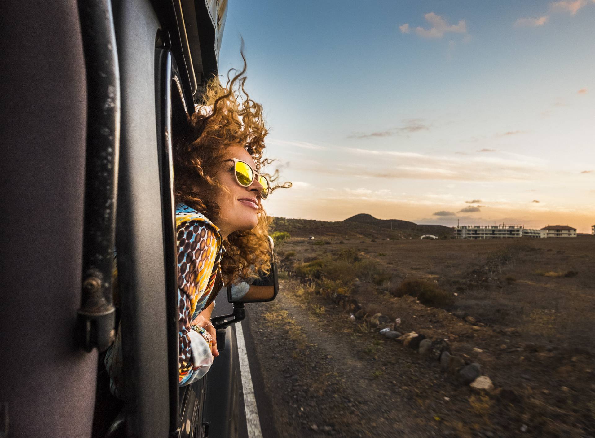 beautiful caucasian young woman travel outside the car with wind in the curly hair, motion and movement on the road discovering new places during a nice sunset, enjoy and joyful freedom concept