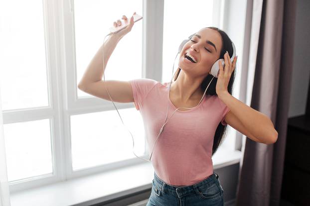Positive young woman stand and listen to music through headphones in room. She dance and enjoy. Model smile. She hold hand on headphone and show piece sign.