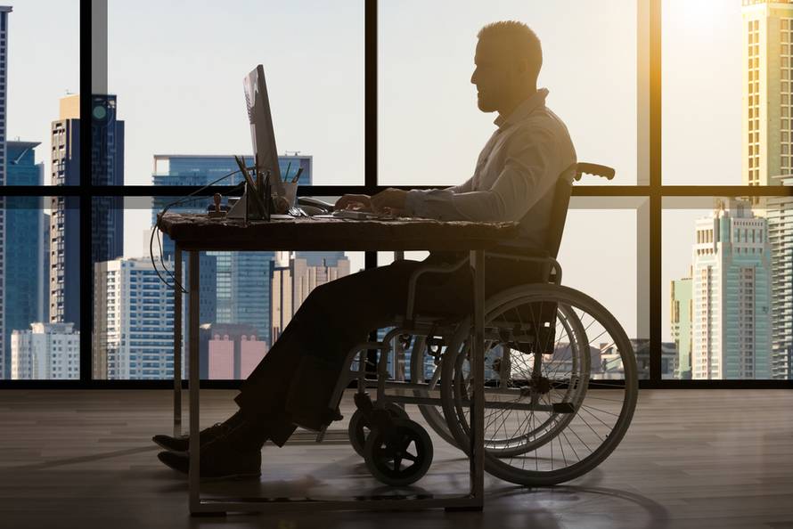 Disabled,Businessman,Sitting,In,Wheelchair,Using,Computer,At,Workplace