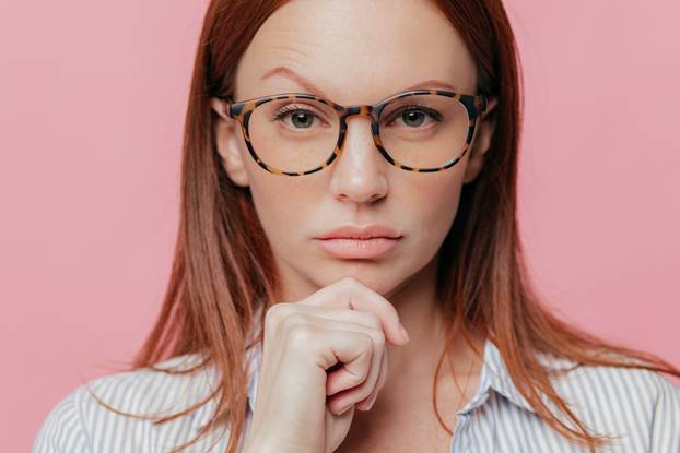 Close up portrait of self confident female business owner holds chin, raises eyebrow, wears optical glasses, has dark hair, dressed in elegant shirt, looks seriously at camera, models in studio