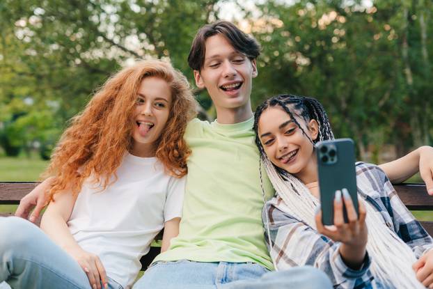 Portrait of a happy three teenagers sitting on the bench and making photo on smartphone. Relaxing on the bench in university campus. Sunny summer day.