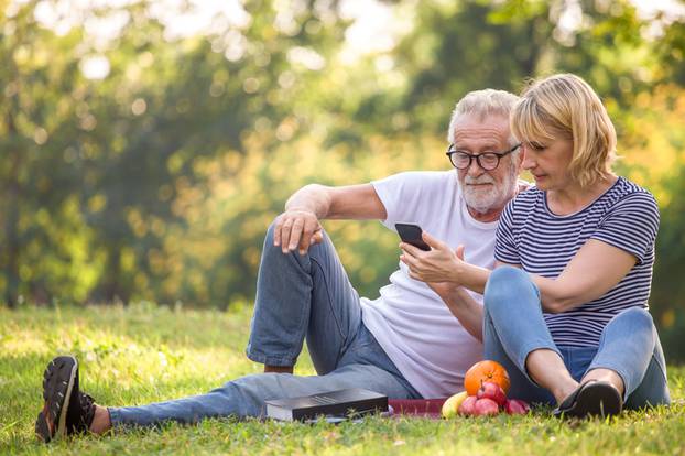 Happy senior couple relaxing in park using smartphone together .