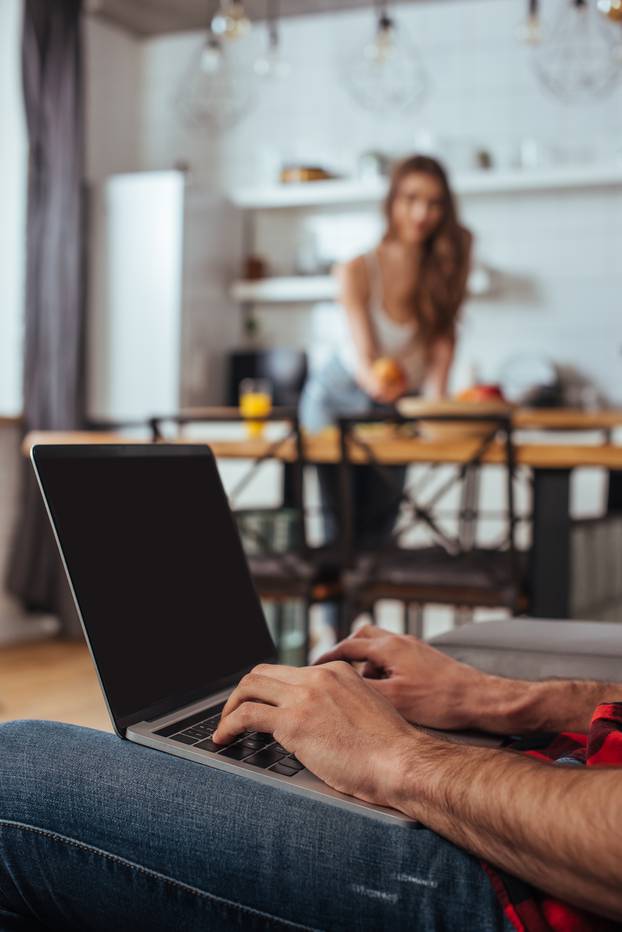 cropped view of freelance typing on laptop with blank screen in kitchen near girlfriend on background