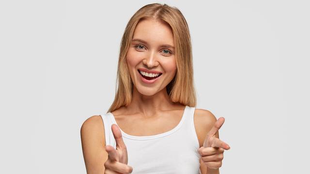 Portrait of happy cute blonde female with joyful expression, points at you with fore fingers directly at camera, chooses companions for entertainment, dressed casually, isolated over white background