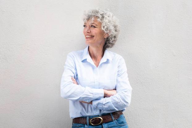 smiling older woman with arms crossed by gray background and looking away
