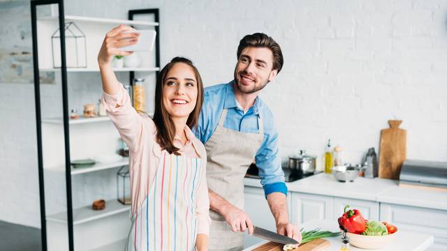 girlfriend taking selfie while boyfriend cutting green onion in kitchen