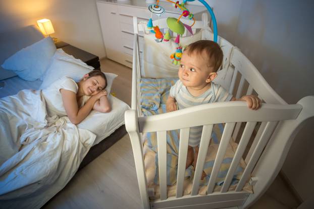 Portrait of baby standing in crib and looking at tired mother th