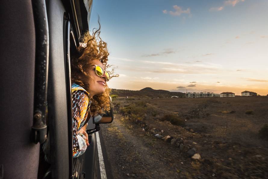 beautiful caucasian young woman travel outside the car with wind in the curly hair, motion and movement on the road discovering new places during a nice sunset, enjoy and joyful freedom concept