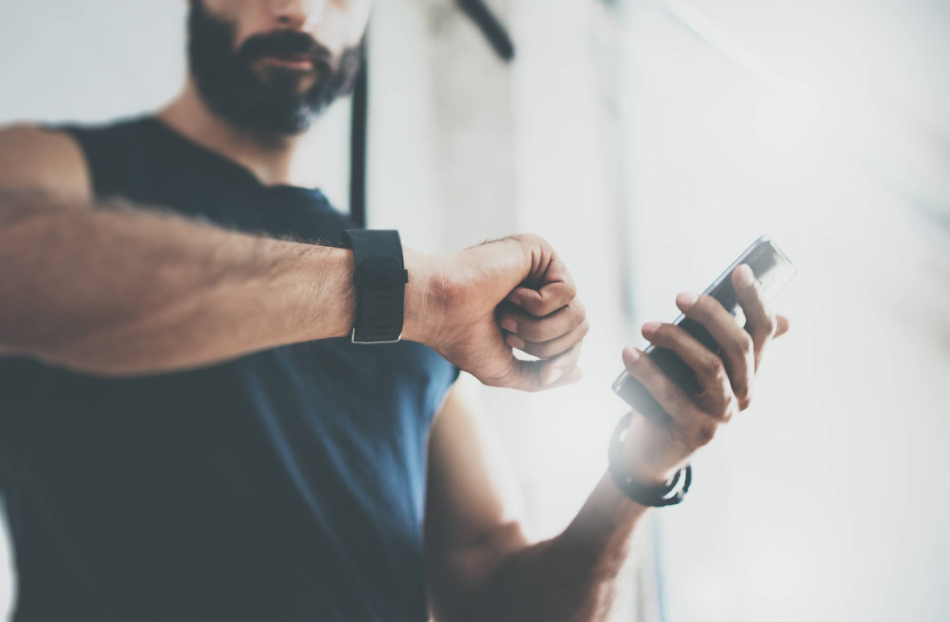 Close-up Shot Bearded Sportive Man After Workout Session Checks Fitness Results Smartphone.Adult Guy Wearing Sport Tracker Wristband Arm.Training hard inside gym.Horizontal bar background.Blurred.