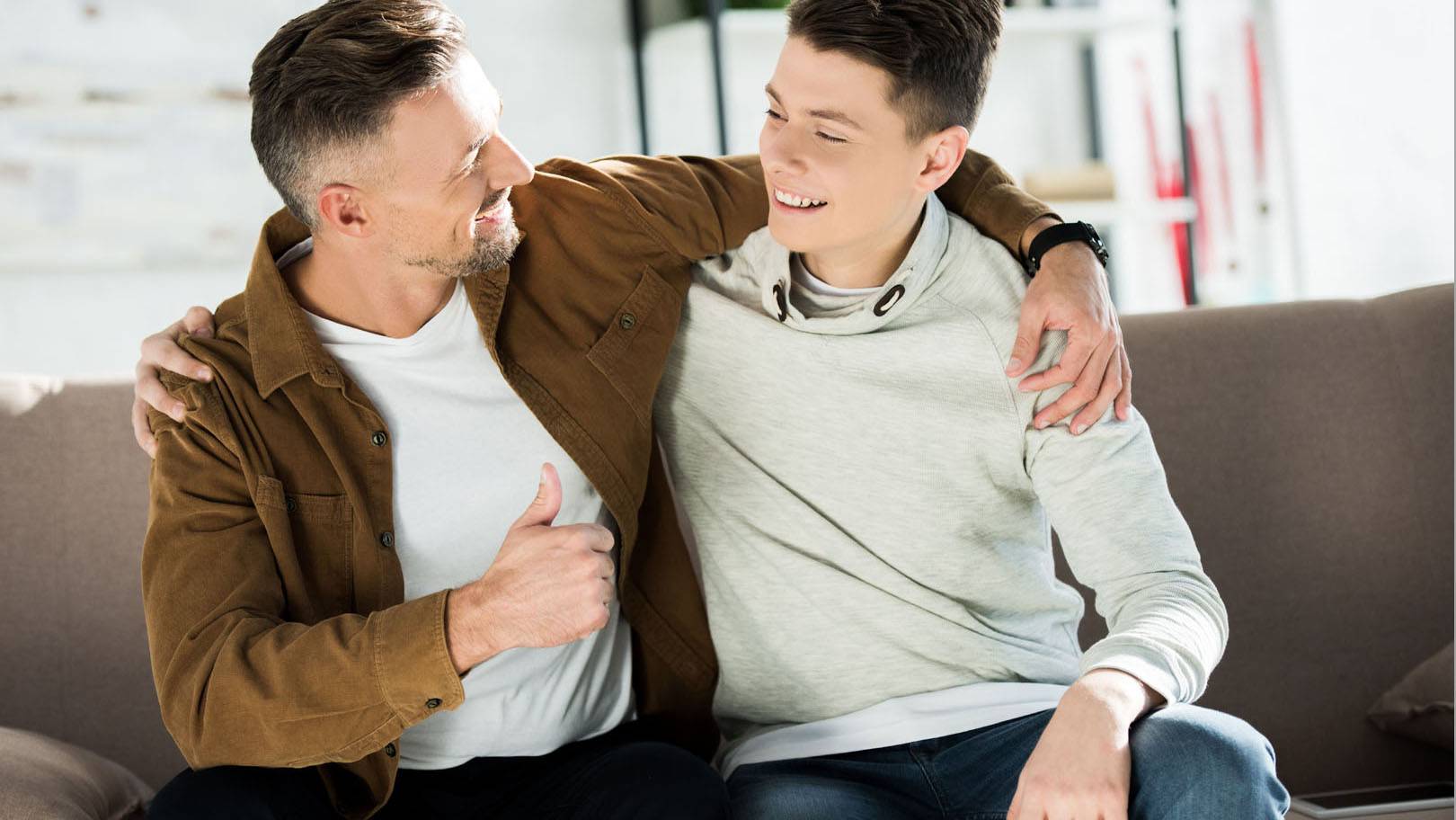 Smiling,Father,And,Teen,Son,Hugging,On,Sofa,At,Home