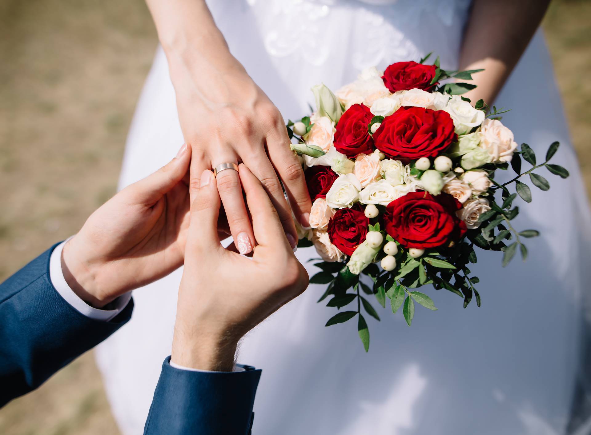 Love and marriage. Fine art rustic wedding ceremony outside. Groom putting golden ring on the bride's finger. Bouquet of red and white roses in hands