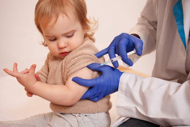 The doctor vaccinates the child against coronavirus. The child is crying and afraid. A man in a robe, hat, mask and gloves makes an injection to the child. Home quarantine