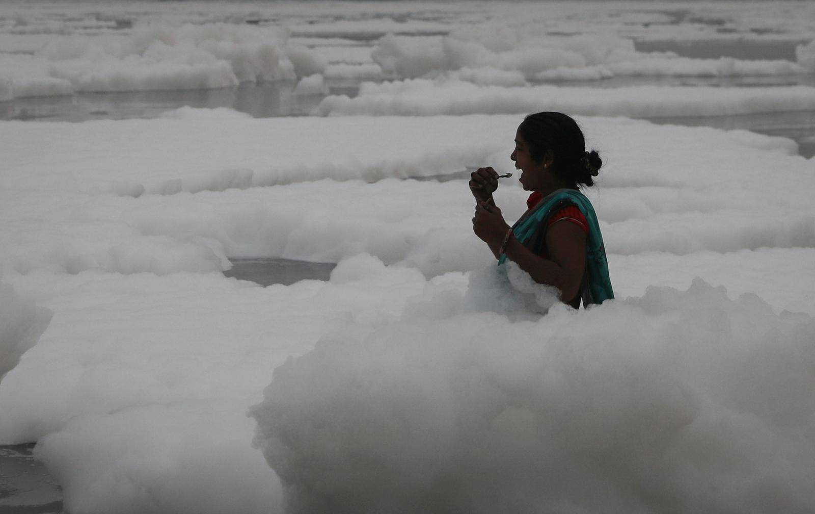 A woman brushes her teeth as she stands amidst the foam covering the polluted Yamuna river on a smoggy morning in New Delhi