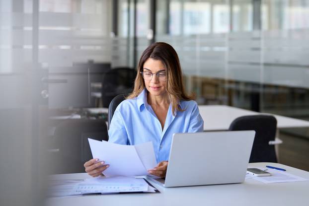 Businesswoman,Of,Middle,Age,Manager,Executive,Reading,Paper,Using,Laptop