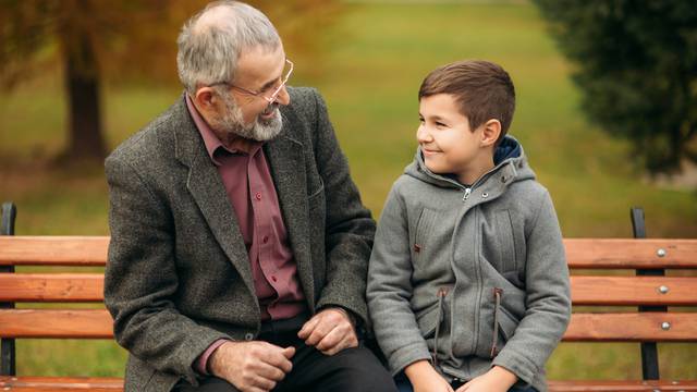 Grandpa and his grandson spend time together in the park. They are sitting on the bench and look to each other