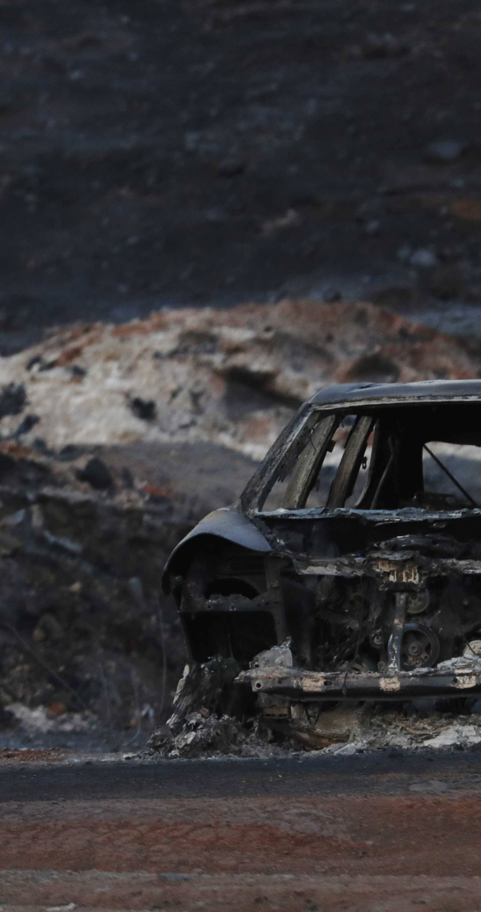 The burnt wreckage of a vehicle is seen along a road as a flames are seen, in the aftermath of the Woolsey fire in Malibu, Southern California