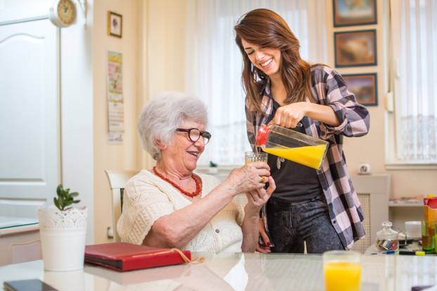 Happy granddaughter pouring orange juice in glass for grandmother in living room.