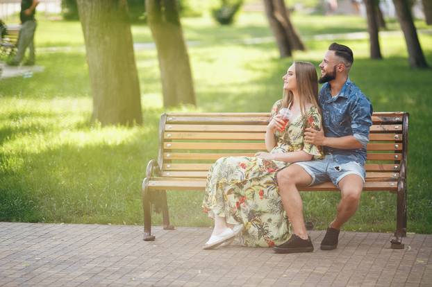 Fashion young teenage couple resting in a city park with drinks sitting on a bench on a sunny summer day