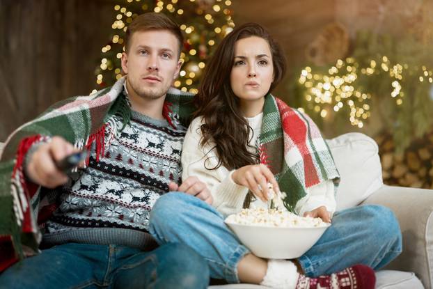 young couple husband and wife wrapped in plaid watching TV eating popcorn on the sofa in room decorated for celebrating new year christmas looking concentrated