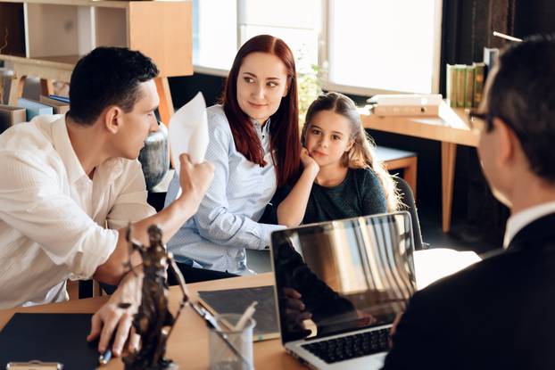 Stunned adult man smashes paper sitting at table next to woman hugging little girl.