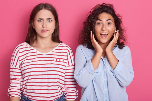 Close up portrait of two stupefied young women standing closely to each other, stare with bugged eyes, being shocked by sudden news or gossip, females posing over pink background. Emotions concept.