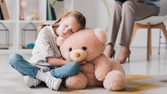 lonely little child with teddy bear sitting on floor with psychologist sitting on background