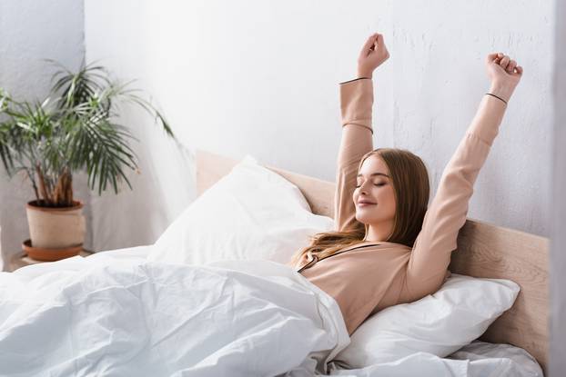 awake and joyful woman in satin pajamas stretching in bed