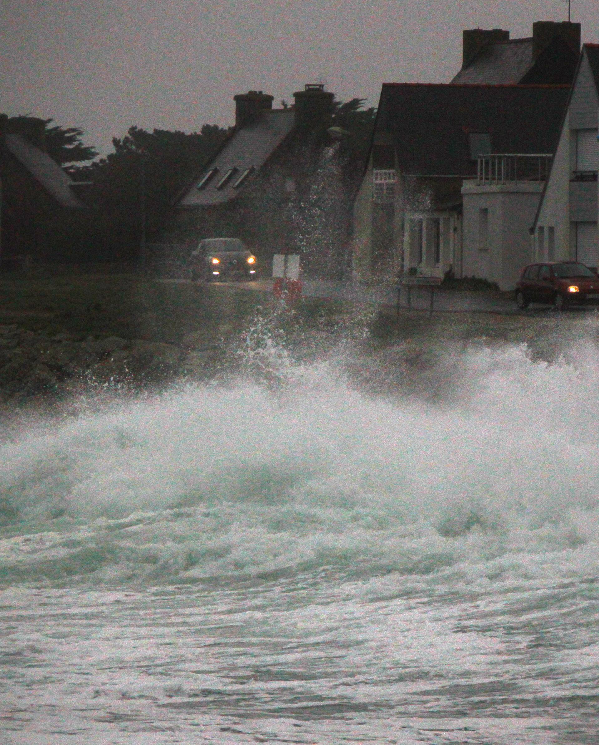 Cars drive past waves breaking on the Brittany coast after storm Eleanor hit Saint-Guenole