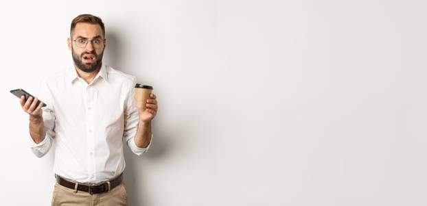 Image of man drinking coffee, feeling confused about strange message on mobile phone, standing over white background