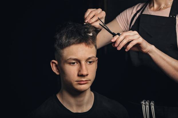 Professional hairdressed. Portrait of handsome man with model haircut. New trendy modern hairstyle. Barber is shearing man hair. Young man in barbershop. Horizontal shot. Black background.