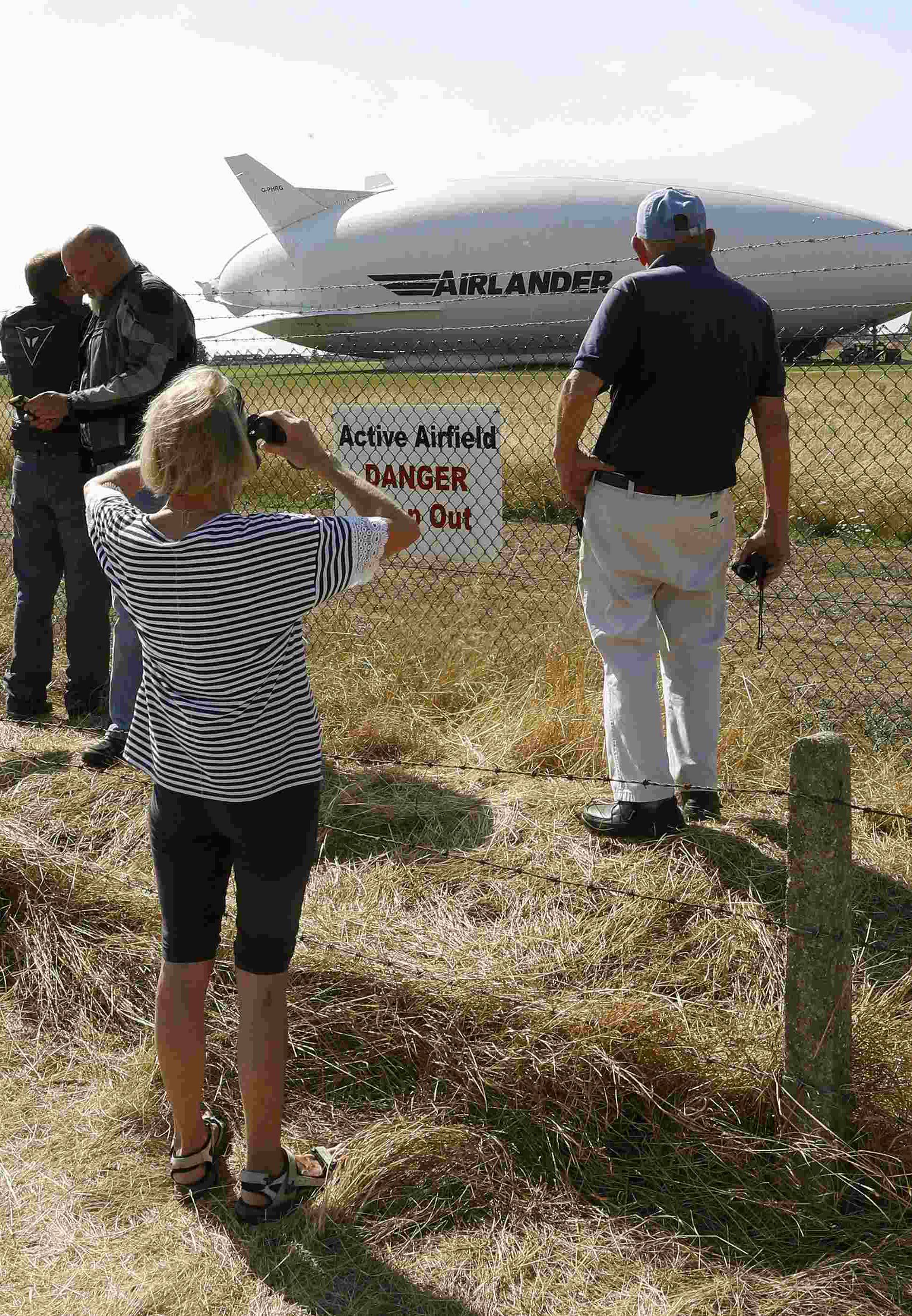 People look through the perimeter fence at the Airlander 10 hybrid airship following a crash-landing during a test flight at Cardington Airfield in Britain