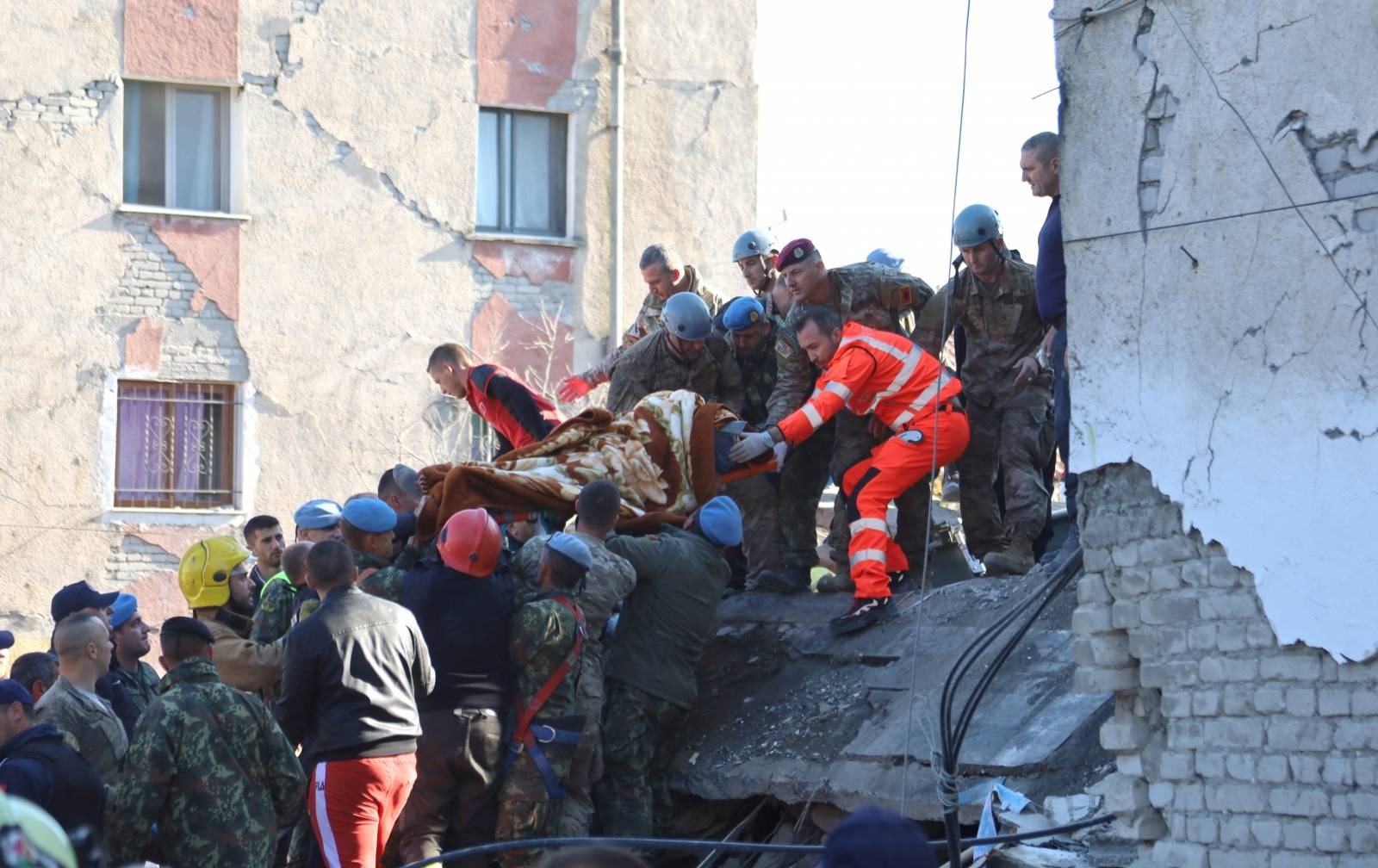 Military and emergency personnel carry an injured man on a stretcher near a damaged building in