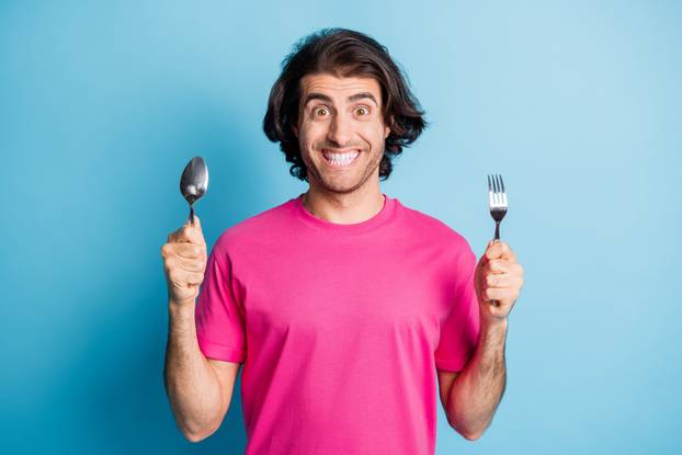 Portrait of nice funny cheerful guy holding in hands fork spoon preparing wait lunch isolated over bright blue color background