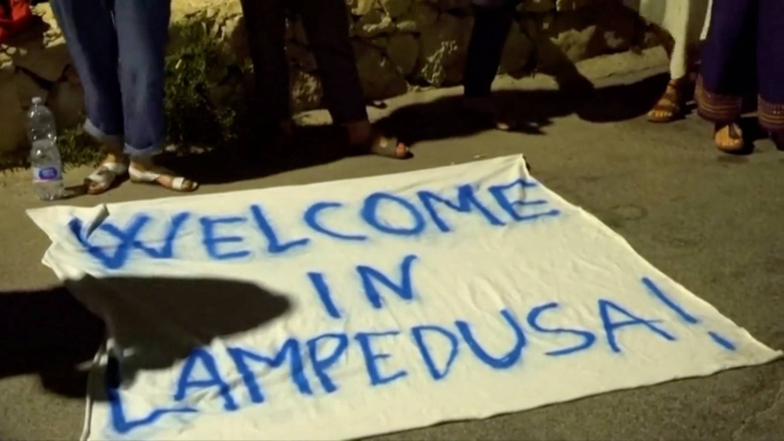 People stand near a banner reading "Welcome in Lampedusa" as migrants disembark a rescue boat in the port of Lampedusa