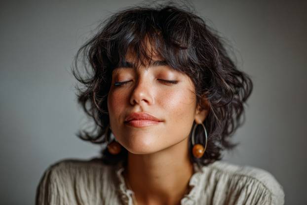 Woman with curly hair and earrings enjoys a moment of tranquility in a softly lit room during a calm afternoon