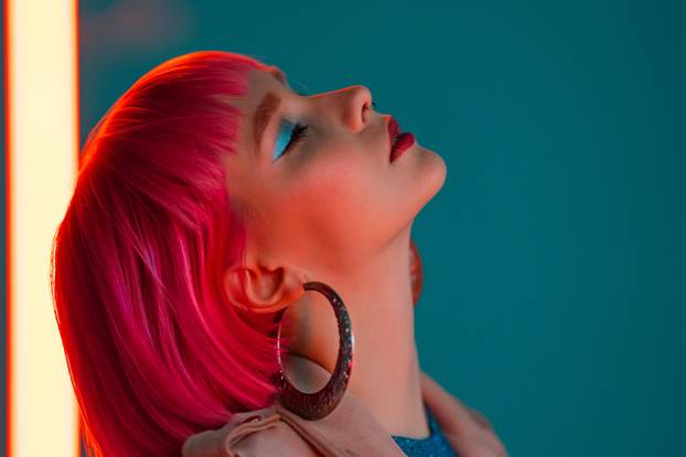 Eccentric woman with pink hair posing near led neon lamp on teal background. Charming odd girl, nightlife concept. Modern model pop outfit, influencer lifestyle.